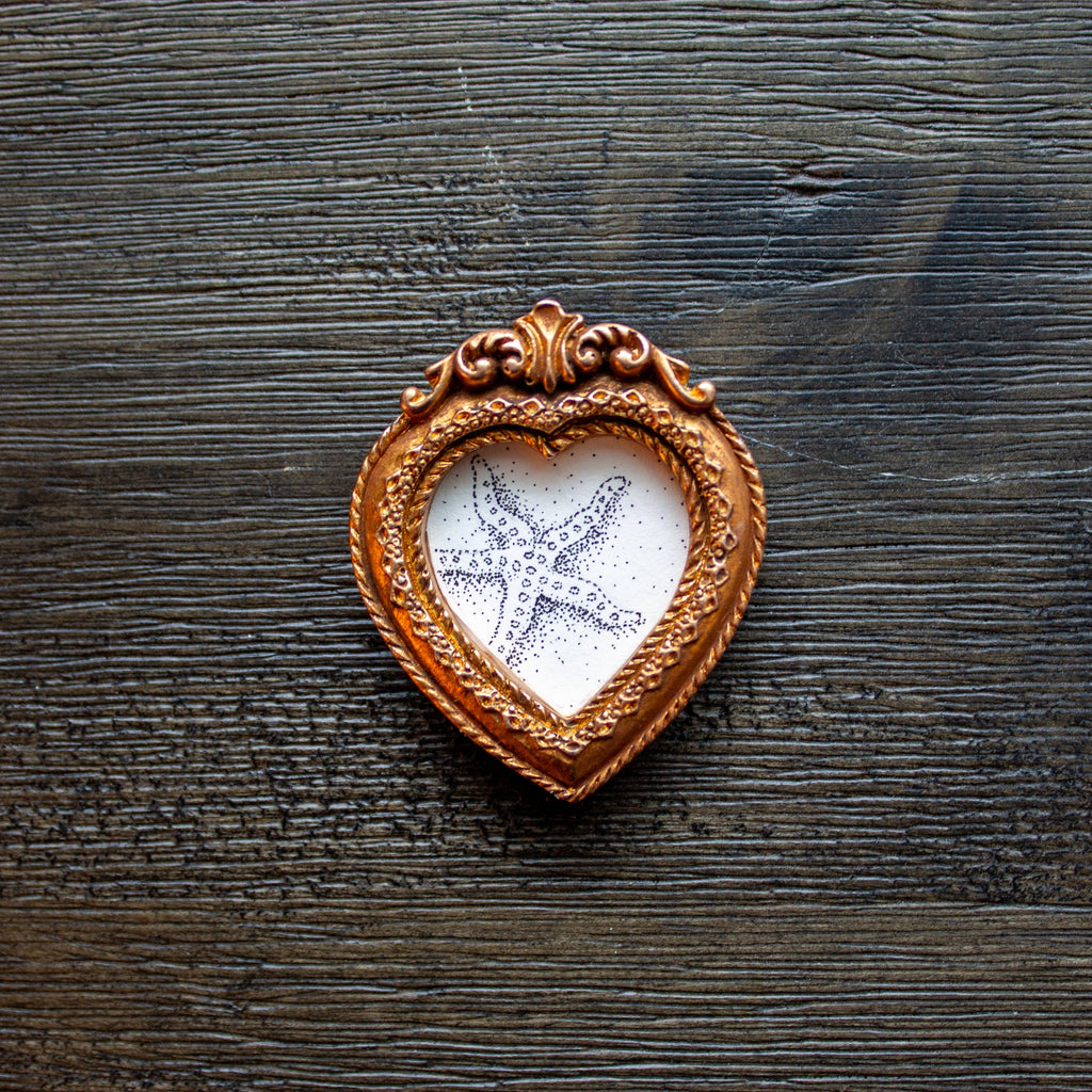 A heart-shaped wooden keepsake frame with an ornate copper-brown border, containing a white textured insert with a printed or painted starfish design, placed on a dark rustic wooden surface.